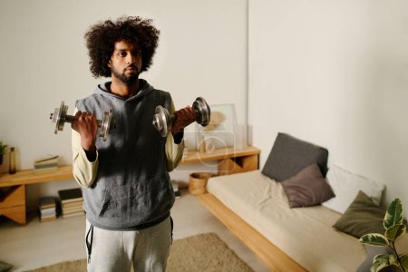 Photo for Young multiethnic man lifting heavy metallic dumbbells while standing in front of camera in bedroom and doing physical exercises - Royalty Free Image