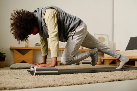 Photo for Young man in sweatpants and hoodie doing press ups while standing on the floor with one leg stretched and the other bent in knee - Royalty Free Image