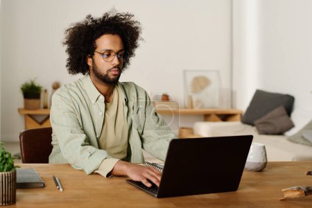 Téléchargez les photos : Jeune freelance masculin multiethnique sérieux ou étudiant regardant écran d'ordinateur portable et dactylographier tout en travaillant sur un nouveau projet en ligne - en image libre de droit