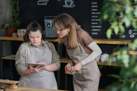 Photo for Young waitress with Down syndrome showing something in tablet to colleague during discussion of working points by counter - Royalty Free Image