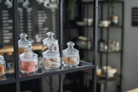 Foto de Grupo de frascos que contienen macarrones y otros dulces de pie en el estante de gran pantalla en la cafetería moderna contra el lugar de trabajo del camarero o empleado - Imagen libre de derechos