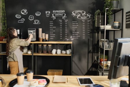 Téléchargez les photos : Jeune femme trisomique en tenue de travail debout à la machine à café et préparant du cappuccino ou une autre boisson aromatique - en image libre de droit
