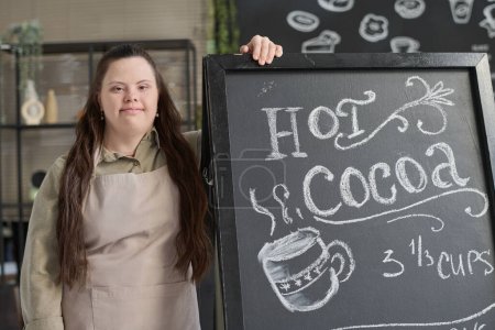 Photo for Young woman with Down syndrome standing by blackboard with special offer written with white chalk and looking at camera - Royalty Free Image