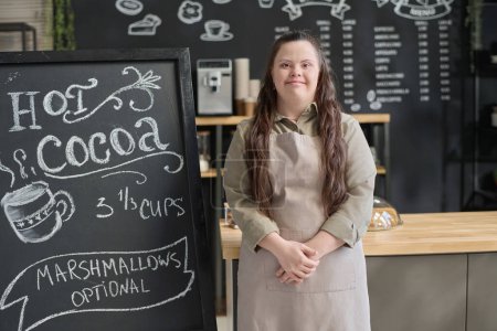 Photo for Smiling waitress with Down syndrome looking at camera while standing by blackboard with special offer of the day written with white chalk - Royalty Free Image