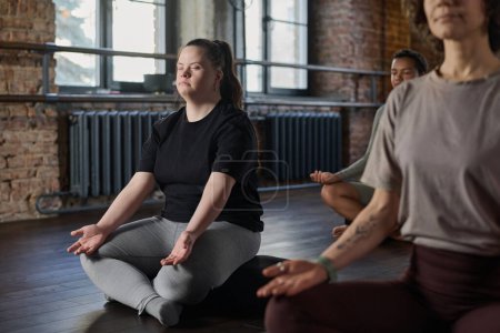 Foto de Mujer joven activa con síndrome de Down sentada en pose de loto con los ojos cerrados y meditando entre otras personas en el gimnasio - Imagen libre de derechos