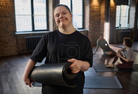 Photo for Young smiling sportswoman with Down syndrome holding rolled mat and looking at camera while standing in gym against two women - Royalty Free Image