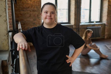 Photo for Young woman with disability keeping hand on metallic bar for exercising and looking at camera while standing against girl sitting on mat - Royalty Free Image