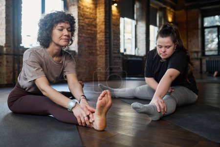 Photo for Girl with Down syndrome repeating after young female fitness trainer while both sitting on mats in gym during physical exercise - Royalty Free Image