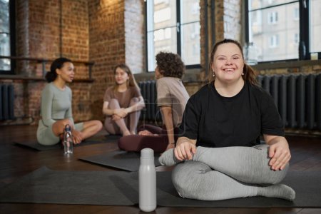 Photo for Young cheerful woman with Down syndrome in activewear sitting on mat and looking at camera against group of active girls in gym - Royalty Free Image