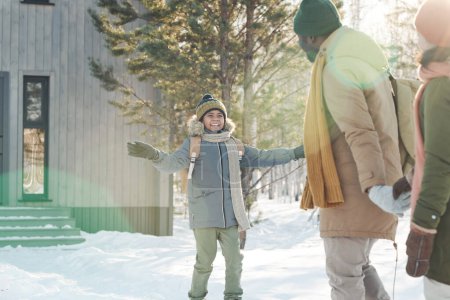 Téléchargez les photos : Joyeux garçon mignon en vêtements d'hiver chauds regardant son père tout en marchant devant ses parents tout en se dirigeant vers leur maison de campagne - en image libre de droit