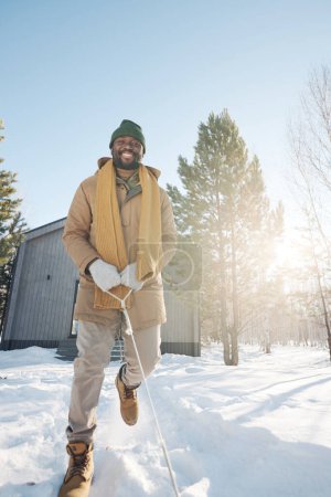 Téléchargez les photos : Jeune homme afro-américain joyeux en vêtements d'hiver tirant la corde de traîneau et descendant le sentier de neige sur une journée d'hiver ensoleillée et givrée - en image libre de droit