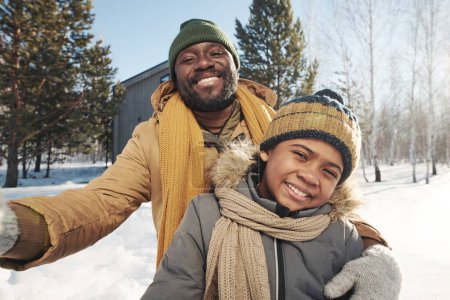 Photo for Cheerful African American father and son in winterwear looking at camera in rural environment on frosty winter day while taking selfie - Royalty Free Image