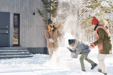 Téléchargez les photos : Joyeux jeune famille interculturelle en vêtements d'hiver jouant avec la neige tout en profitant des vacances d'hiver dans un environnement naturel par maison de campagne - en image libre de droit