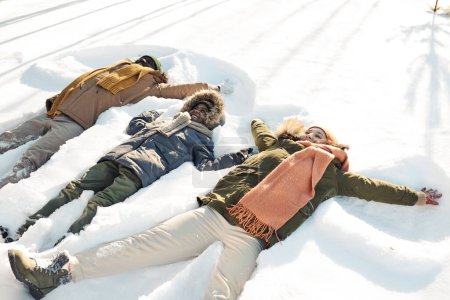 Foto de Tres alegres personas interculturales en ropa de invierno tumbadas en la nieve mientras juegan y se divierten en el ocio o durante las vacaciones de invierno - Imagen libre de derechos