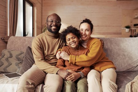Photo for Happy young man and woman in casualwear embracing their adorable son while sitting on couch in living room in cottage and looking at camera - Royalty Free Image