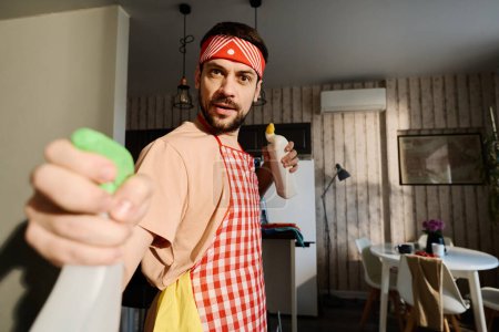 Foto de Tipo en delantal sosteniendo detergentes y rociando uno de ellos durante las tareas domésticas mientras está de pie delante de la cámara - Imagen libre de derechos