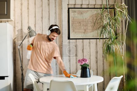 Photo for Young man in headphones, casualwear and rubber gloves wiping kitchen table and spraying detergent while carrying out domestic chores - Royalty Free Image