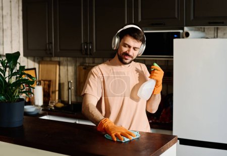 Foto de Chico feliz con plumero y detergente escuchando música en los auriculares y limpiando el mostrador de la cocina durante las tareas domésticas - Imagen libre de derechos