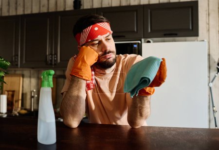 Foto de Joven hombre cansado en diadema y camiseta mirando el plumero azul mientras está de pie junto al mostrador de la cocina con botella de plástico de detergente - Imagen libre de derechos