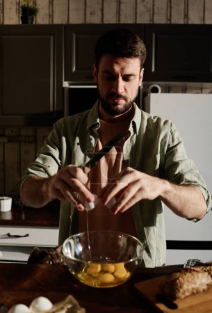 Photo for Bearded guy breaking several eggs into glass bowl while standing in front of camera and preparing omelet by kitchen table or counter - Royalty Free Image