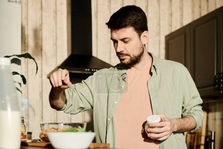 Téléchargez les photos : Guy saupoudrer de sel de mer dans un bol avec des œufs crus cassés tout en se tenant devant la caméra dans la cuisine et préparer le petit déjeuner - en image libre de droit
