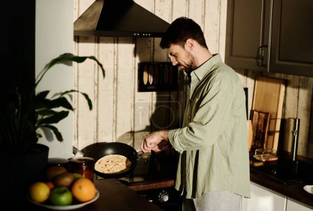 Photo for Side view of young man in grey cotton shirt holding hot frying pan while standing by electric stove and cooking omelet for breakfast - Royalty Free Image