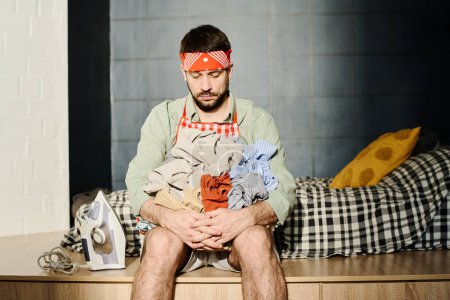 Téléchargez les photos : Jeune homme fatigué avec une pile de vêtements froissés dans les mains assis sur le bord de son lit devant la caméra et en les regardant - en image libre de droit