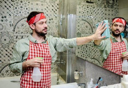Téléchargez les photos : Guy dans le bandeau et tablier pulvérisation de détergent sur le miroir dans la salle de bain et essuyer avec chiffon humide pendant les tâches ménagères quotidiennes - en image libre de droit