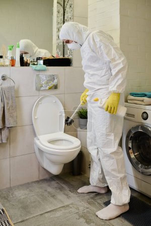 Photo for Young man in respirator, protective gloves and hazmat suit standing in front of ceramic toilet bowl and cleaning it with detergent and brush - Royalty Free Image