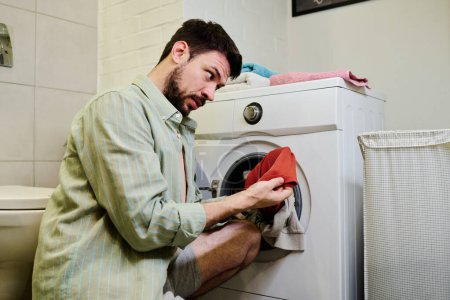 Téléchargez les photos : Homme perplexe en chemise grise assis sur des squats devant la machine à laver et regardant la serviette rouge tirée de tas d'autres vêtements - en image libre de droit