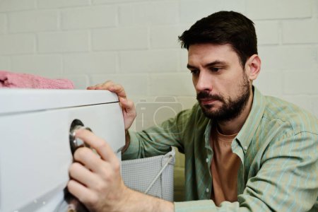Foto de Hombre joven serio en cuclillas por lavadora automática en el baño y la elección de los ajustes antes de pulsar el botón de inicio - Imagen libre de derechos