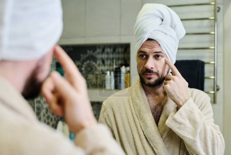Photo for Young bearded man with towel on head putting moisturizing patches on under eye area while having beauty procedure after morning shower - Royalty Free Image