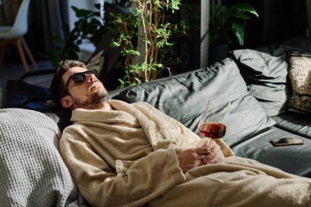 Foto de Joven en albornoz y gafas de sol soñando despierto con una copa de vino tinto en la mano mientras se relaja en el sofá con almohadas - Imagen libre de derechos