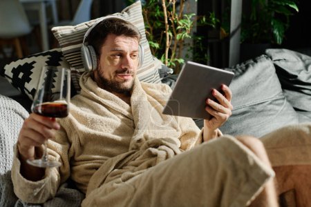 Téléchargez les photos : Jeune homme détendu avec un verre de vin regardant l'écran de la tablette tout en étant allongé sur le canapé et en regardant un film en ligne ou en communiquant par chat vidéo - en image libre de droit