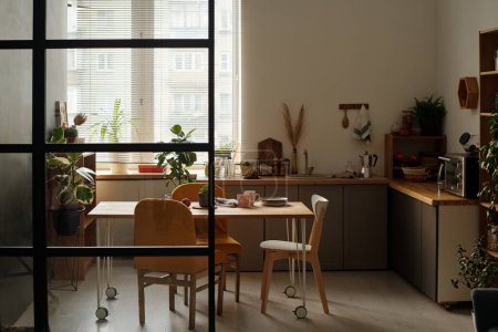 Foto de Parte de la amplia cocina con mostrador, grupo de sillas, mesa de madera con utensilios de cocina y plantas domésticas verdes que crecen en macetas - Imagen libre de derechos