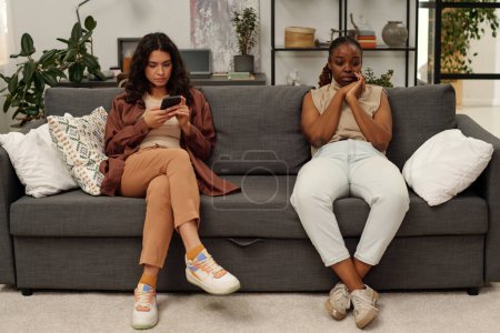 Foto de Joven mujer celosa desplazándose en el teléfono inteligente de la novia afroamericana mientras ambos están sentados en el sofá en la sala de estar durante el conflicto - Imagen libre de derechos