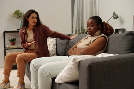 Foto de Mujer afroamericana trastornada con los brazos cruzados contra el pecho sentada en el sofá junto a su novia nerviosa durante la discusión - Imagen libre de derechos