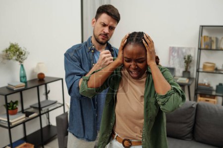 Foto de Joven mujer afroamericana tocando la cabeza con las manos mientras está de pie frente a su marido furioso tratando de golpearla durante la pelea - Imagen libre de derechos
