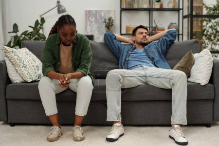 Téléchargez les photos : Jeune femme afro-américaine bouleversée assise sur le canapé à côté de son mari indifférent regardant de côté pendant la querelle - en image libre de droit