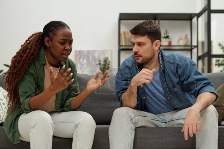 Téléchargez les photos : Jeune femme afro-américaine essayant d'expliquer quelque chose à son mari pendant la dispute alors que les deux assis sur le canapé - en image libre de droit