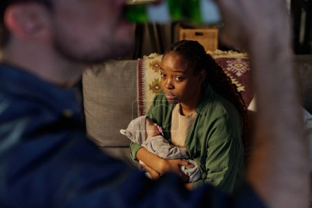 Téléchargez les photos : Focus sur jeune femme afro-américaine contrariée et agacée avec bébé sur les mains en regardant son mari boire de la bière au biberon - en image libre de droit