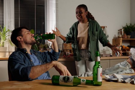 Foto de Joven mujer afroamericana molesta gritando a su marido sentado junto a la mesa de la cocina y bebiendo cerveza de la botella durante el conflicto - Imagen libre de derechos