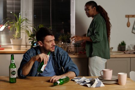 Téléchargez les photos : Jeune homme stressé avec bouteille de bière à la main assis à table dans la cuisine et regardant femme en colère et agacée - en image libre de droit