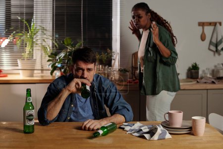 Téléchargez les photos : Homme malheureux avec dépendance à l'alcool assis à table dans la cuisine et boire de la bière contre femme irritée criant sur lui pendant le conflit - en image libre de droit