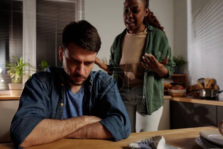 Foto de Joven hombre estresado con los brazos cruzados por el pecho sentado en la mesa de la cocina mientras su esposa irritada gritándole durante la discusión - Imagen libre de derechos