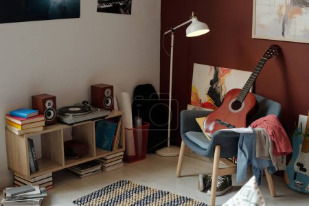 Photo for New acoustic guitar standing on armchair with pile of clothes in front of record player surrounded by books and two sound speakers - Royalty Free Image