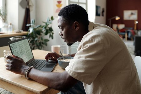 Foto de Joven hombre de negocios sentado a la mesa frente a la pantalla del ordenador portátil mientras desayuna y mira a través de noticias en línea o análisis de datos - Imagen libre de derechos