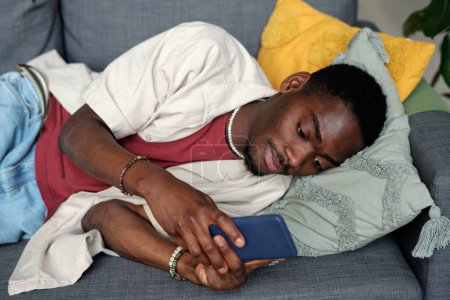 Téléchargez les photos : Jeune homme afro-américain se détendre sur le canapé et regarder à travers les messages en ligne sur l'écran du smartphone ou regarder la vidéo - en image libre de droit