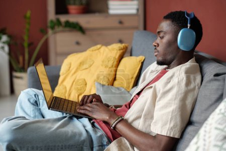 Téléchargez les photos : Jeune homme d'affaires ou blogueur dans un casque regardant l'écran de l'ordinateur portable et tapant tout en étant assis sur le canapé dans le salon pendant le réseau - en image libre de droit