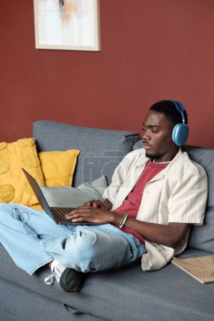 Téléchargez les photos : Jeune homme d'affaires afro-américain moderne en casual et écouteurs assis sur le canapé et l'analyse des données en ligne - en image libre de droit